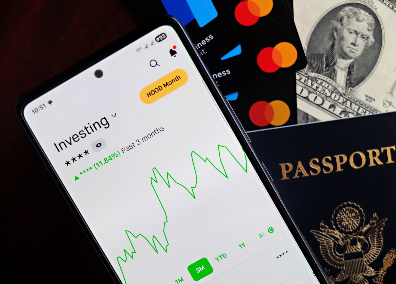 home-hero-bg Smartphone displaying investing app, with credit cards, cash, and passport nearby, symbolizing finance