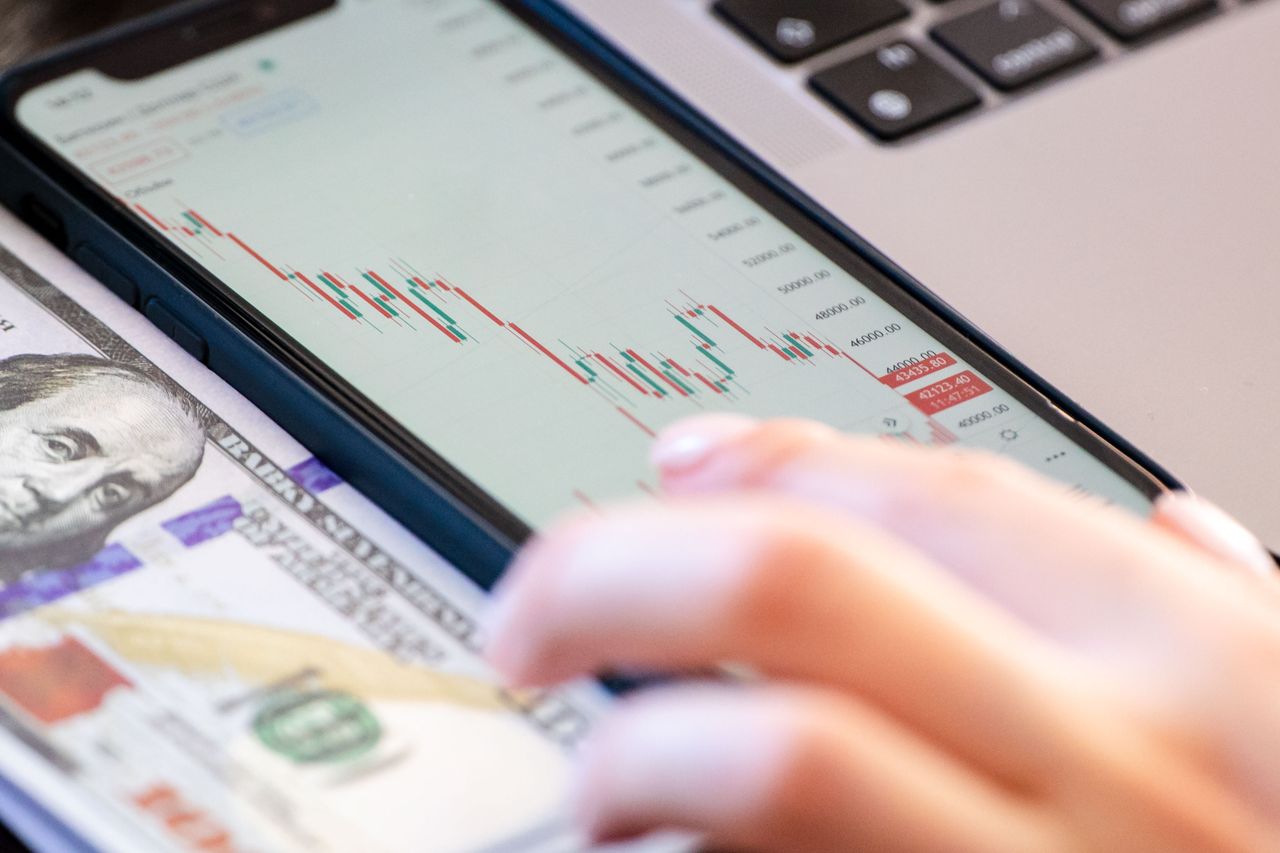 about-01 Close-up of a smartphone displaying stock market data over a dollar bill on a desk.